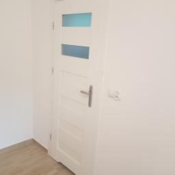 Usługi remontowo budowlane - Białe drzwi wewnętrzne z trzema przeszkleniami w kolorze błękitnym, zamontowane w świeżo wyremontowanym pomieszczeniu z jasnymi ścianami i podłogą wyłożoną płytkami imitującymi drewno.