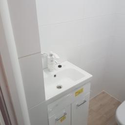 Usługi remontowo budowlane - Biała umywalka z szafką i elektryczną baterią podgrzewającą wodę, zamontowana w łazience wyłożonej białymi płytkami, widok z boku, Serock.