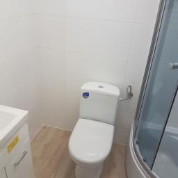 Usługi remontowo budowlane - Nowoczesna, jasna łazienka z białą ceramiką: toaleta, umywalka z szafką i kabina prysznicowa z szarą szybą, wykończona jasnymi płytkami na ścianach i podłodze.