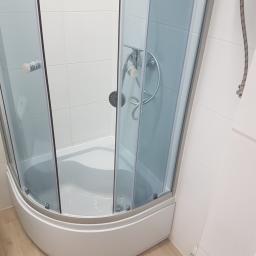 Usługi remontowo budowlane - Narożna kabina prysznicowa z przezroczystymi drzwiami i chromowaną armaturą, na tle białych płytek ściennych i podłogi imitującej drewno.