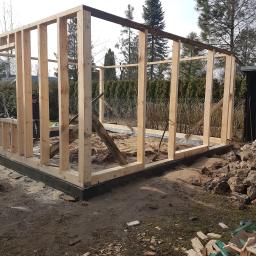 Usługi remontowo budowlane - Konstrukcja szkieletowa budynku w trakcie budowy, widoczne drewniane słupy i belki, fundament z izolacją, teren budowy z piaskiem i resztkami drewna