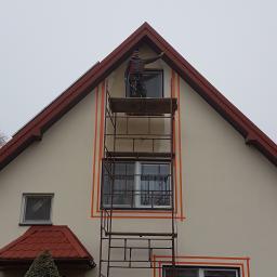 Usługi remontowo budowlane - Pracownik na rusztowaniu maluje elewację domu jednorodzinnego, zabezpieczoną pomarańczową taśmą malarską wokół okien i dachu, w pochmurny dzień.