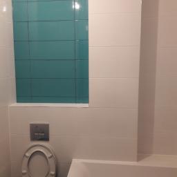 Usługi remontowo budowlane - Wykończona łazienka z białymi i turkusowymi płytkami, widoczna toaleta z otwartą klapą i fragment wanny.