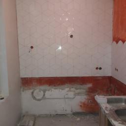 Usługi remontowo budowlane - Ściana w trakcie układania białych, sześciennych płytek w łazience, widoczne otwory na gniazdka elektryczne, częściowo pomalowana ściana na czerwono, na podłodze wiadro i worek z zaprawą.