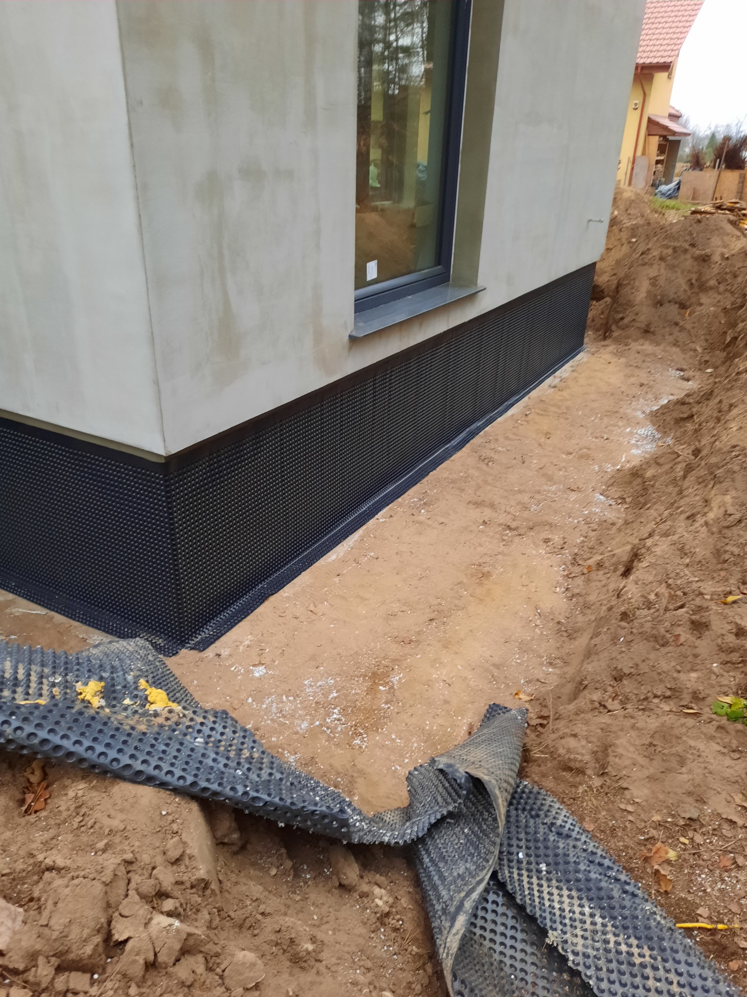 Częściowo zabezpieczone fundamenty budynku mieszkalnego czarną folią izolacyjną z wytłoczeniami, wykop wokół fundamentów, okno z szarą ramą, ślady białego cementu na ziemi.