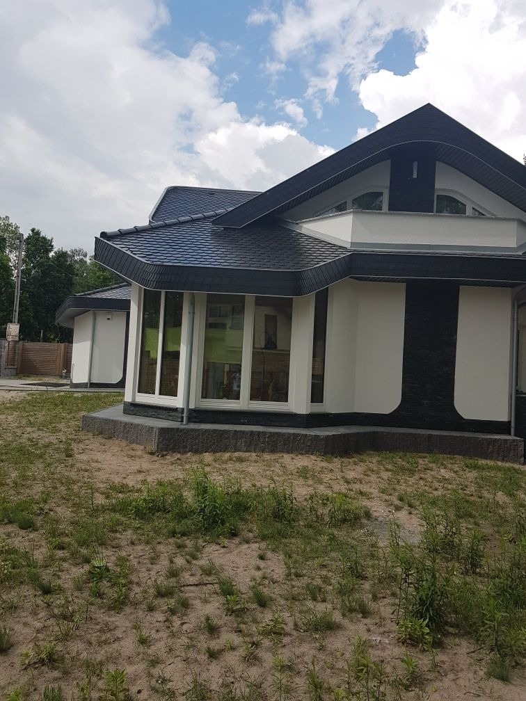 Nowoczesny dom jednorodzinny z czarnym dachem z dachówki bitumicznej i białymi ścianami z czarnymi akcentami, otoczony piaszczystym terenem z młodą zielenią.