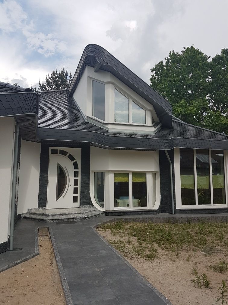 Dom jednorodzinny z dachem pokrytym ciemną dachówką łuskową, białą stolarką okienną o nietypowych kształtach i ścieżką z szarych płyt prowadzącą do wejścia.
