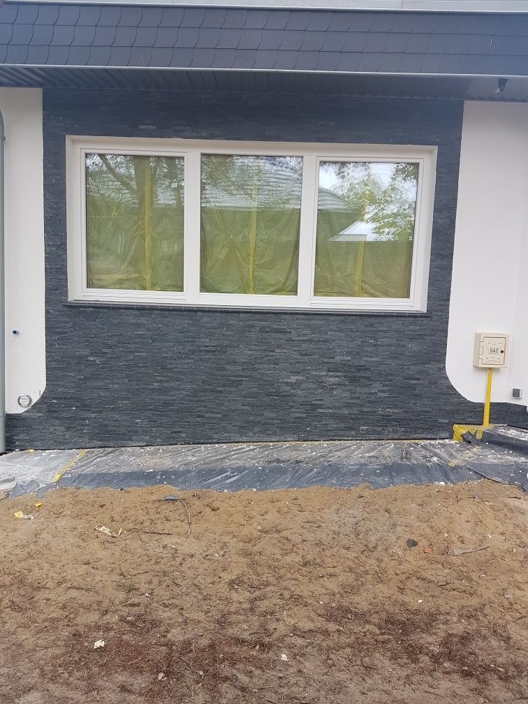 Elewacja budynku z oknem zabezpieczonym folią, wykończona szarą okładziną imitującą kamień, z widoczną skrzynką gazową i przygotowanym podłożem z piasku.