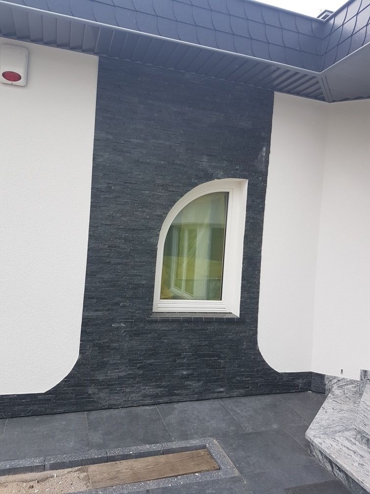 Fragment elewacji domu z oknem o nietypowym, zaokrąglonym kształcie, wykończonej czarną, łupaną okładziną kamienną, kontrastującą z białą, teksturowaną ścianą. Pod oknem widoczny fragment szarego...