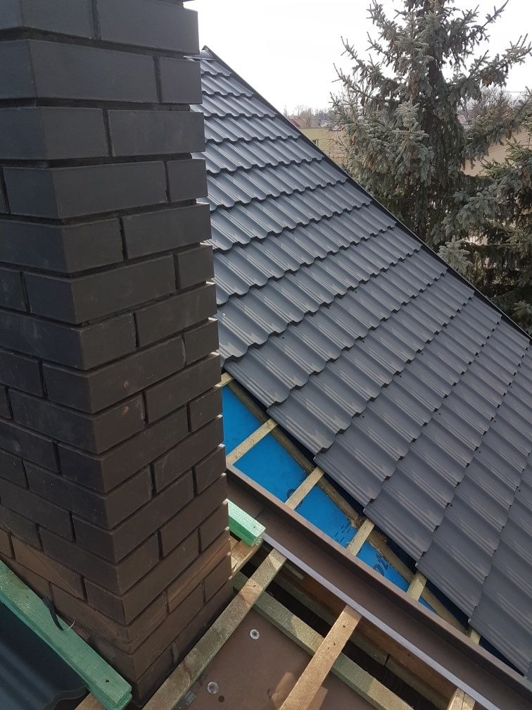 Montaż grafitowej blachodachówki trapezowej przy kominie z ciemnej cegły, widoczne drewniane łaty i niebieska membrana dachowa.