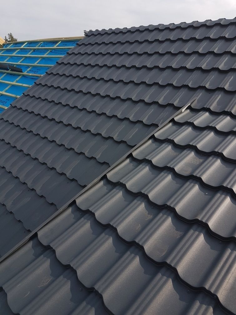 Fragment dachu pokrytego ciemnoszarą blachodachówką, widoczne elementy konstrukcji dachu z membraną w tle.