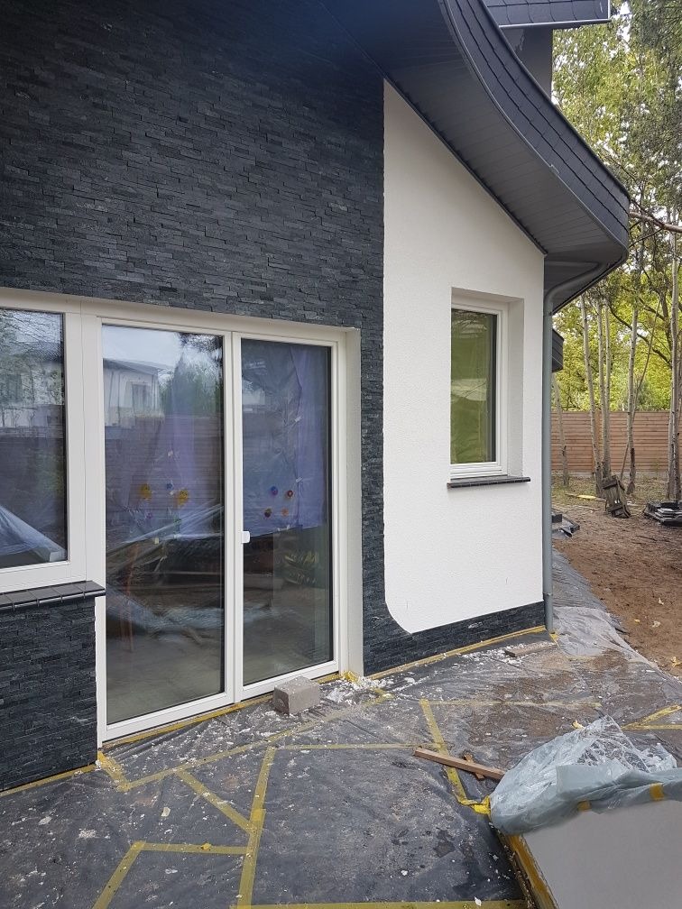 Fragment elewacji domu z wykończeniem z czarnego kamienia dekoracyjnego wokół okien i drzwi przesuwnych, zabezpieczona folią malarską podczas prac wykończeniowych.