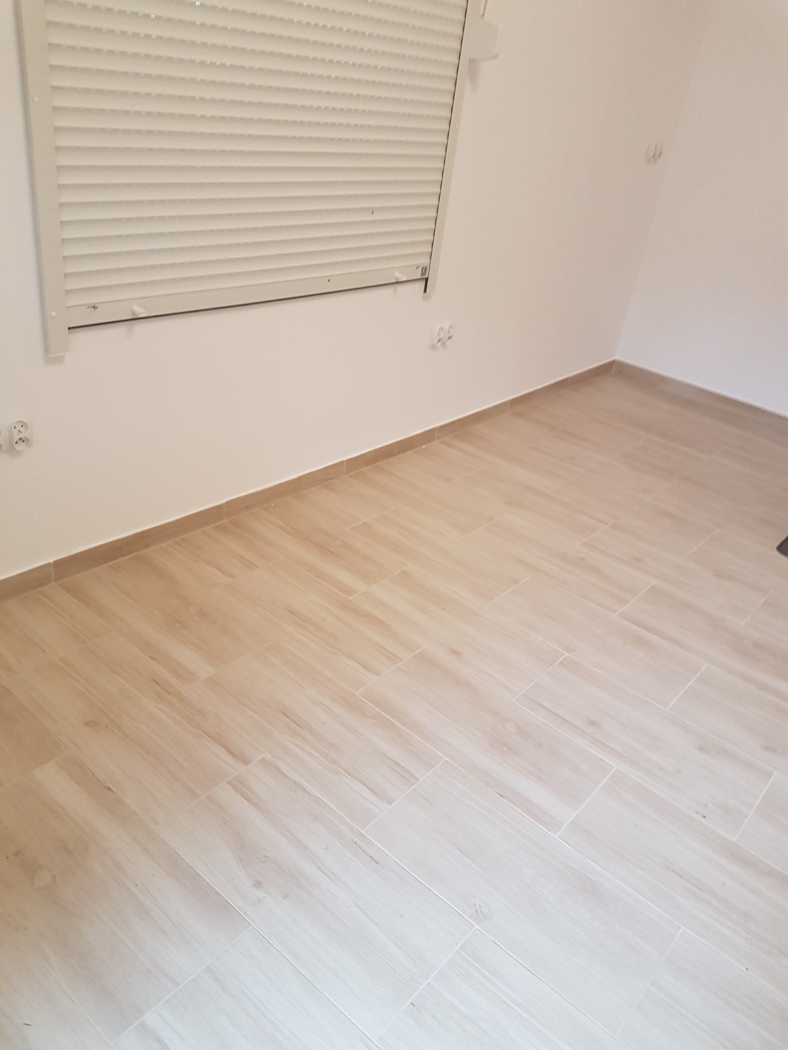 Jasne, drewnopodobne płytki podłogowe ułożone w pustym pokoju z białymi ścianami, widoczne gniazdka elektryczne i okno z żaluzjami.