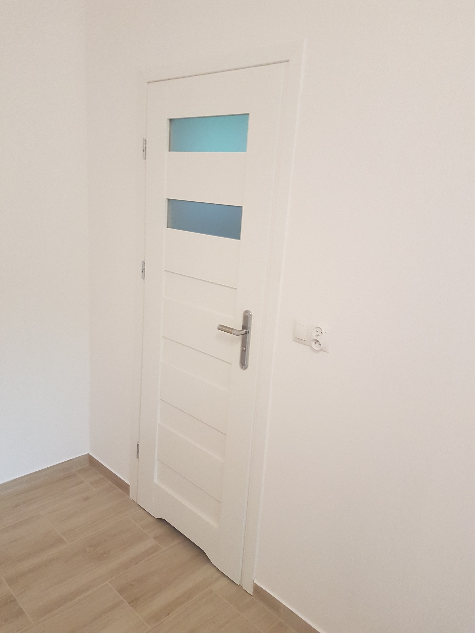 Białe drzwi wewnętrzne z trzema przeszkleniami w kolorze błękitnym, zamontowane w świeżo wyremontowanym pomieszczeniu z jasnymi ścianami i podłogą wyłożoną płytkami imitującymi drewno.