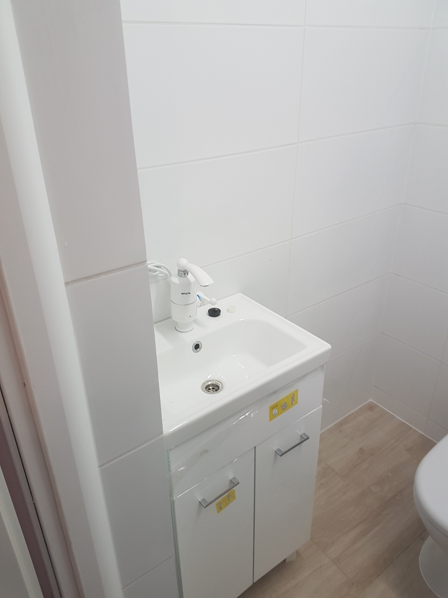 Biała umywalka z szafką i elektryczną baterią podgrzewającą wodę, zamontowana w łazience wyłożonej białymi płytkami, widok z boku, Serock.