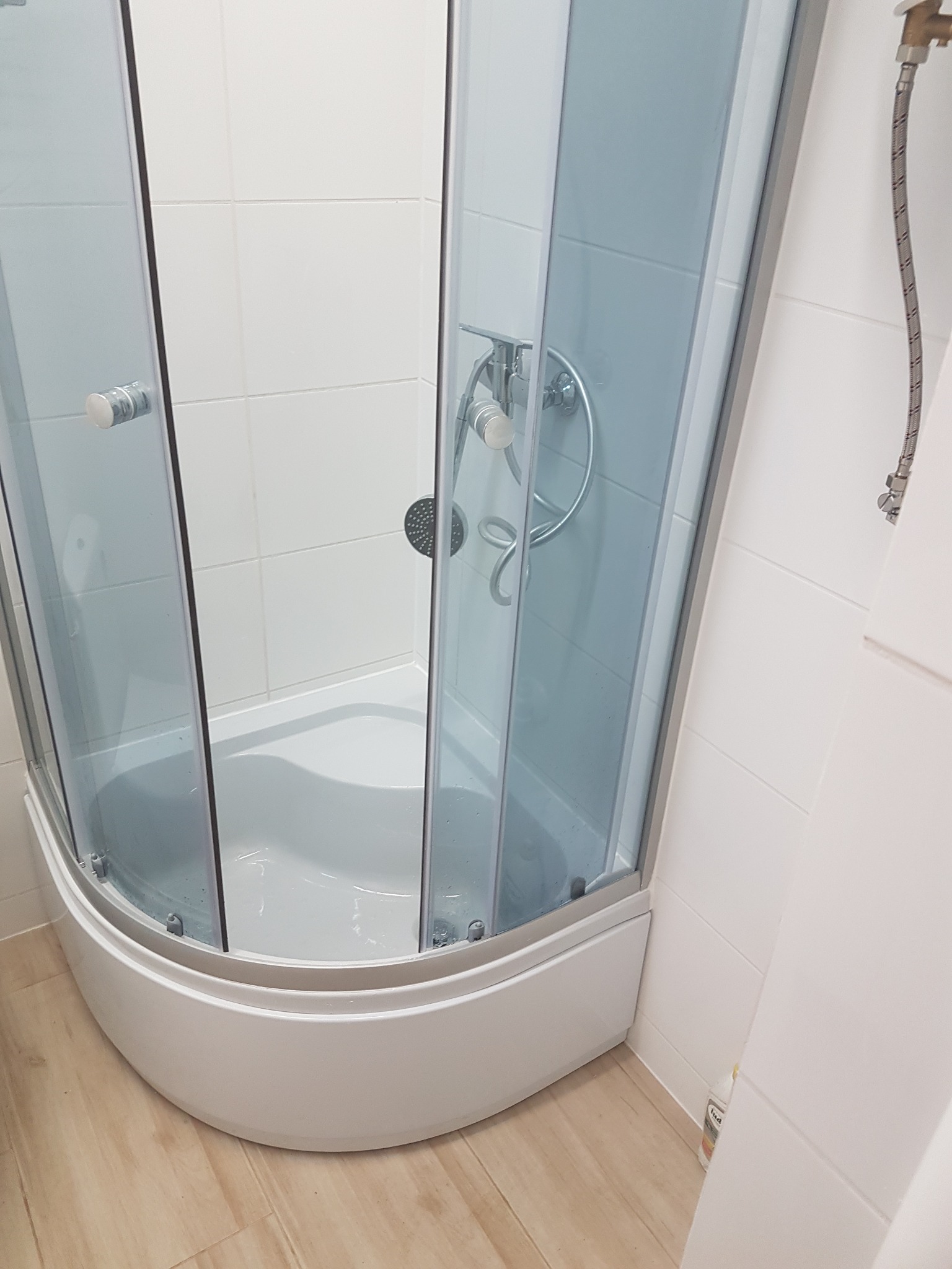 Narożna kabina prysznicowa z przezroczystymi drzwiami i chromowaną armaturą, na tle białych płytek ściennych i podłogi imitującej drewno.