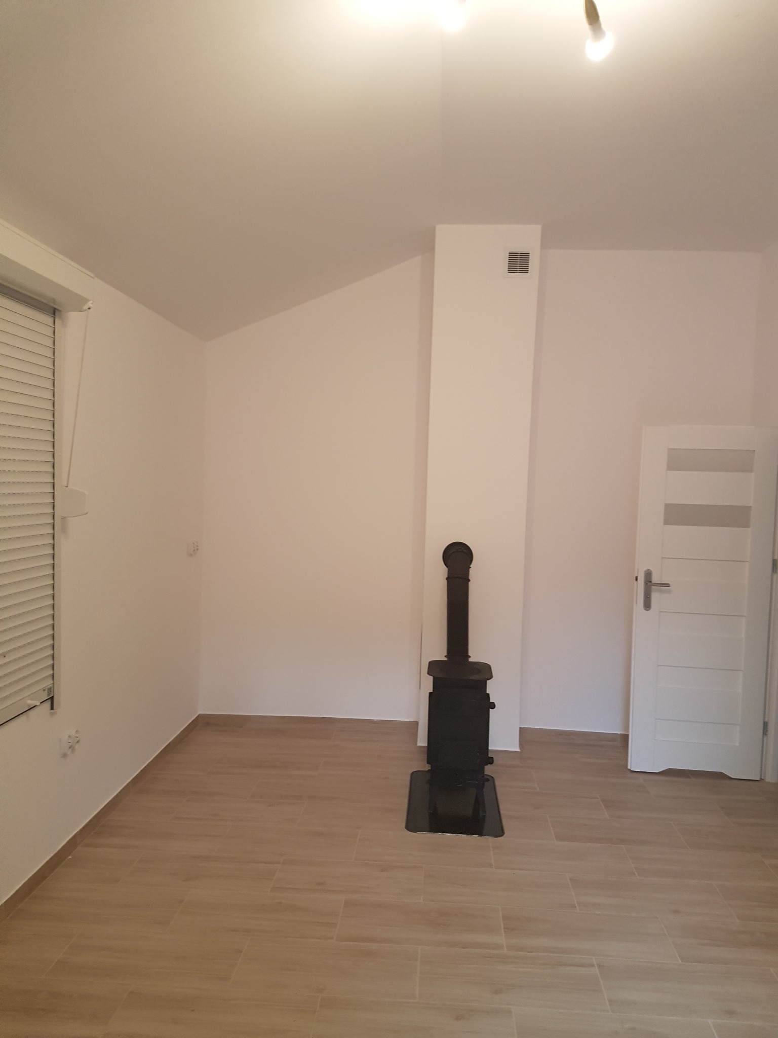 Wnętrze pokoju z ukośnym sufitem, piecem wolnostojącym typu koza z podłączeniem do komina, jasną podłogą z płytek imitujących drewno, białymi ścianami i drzwiami z przeszkleniami.