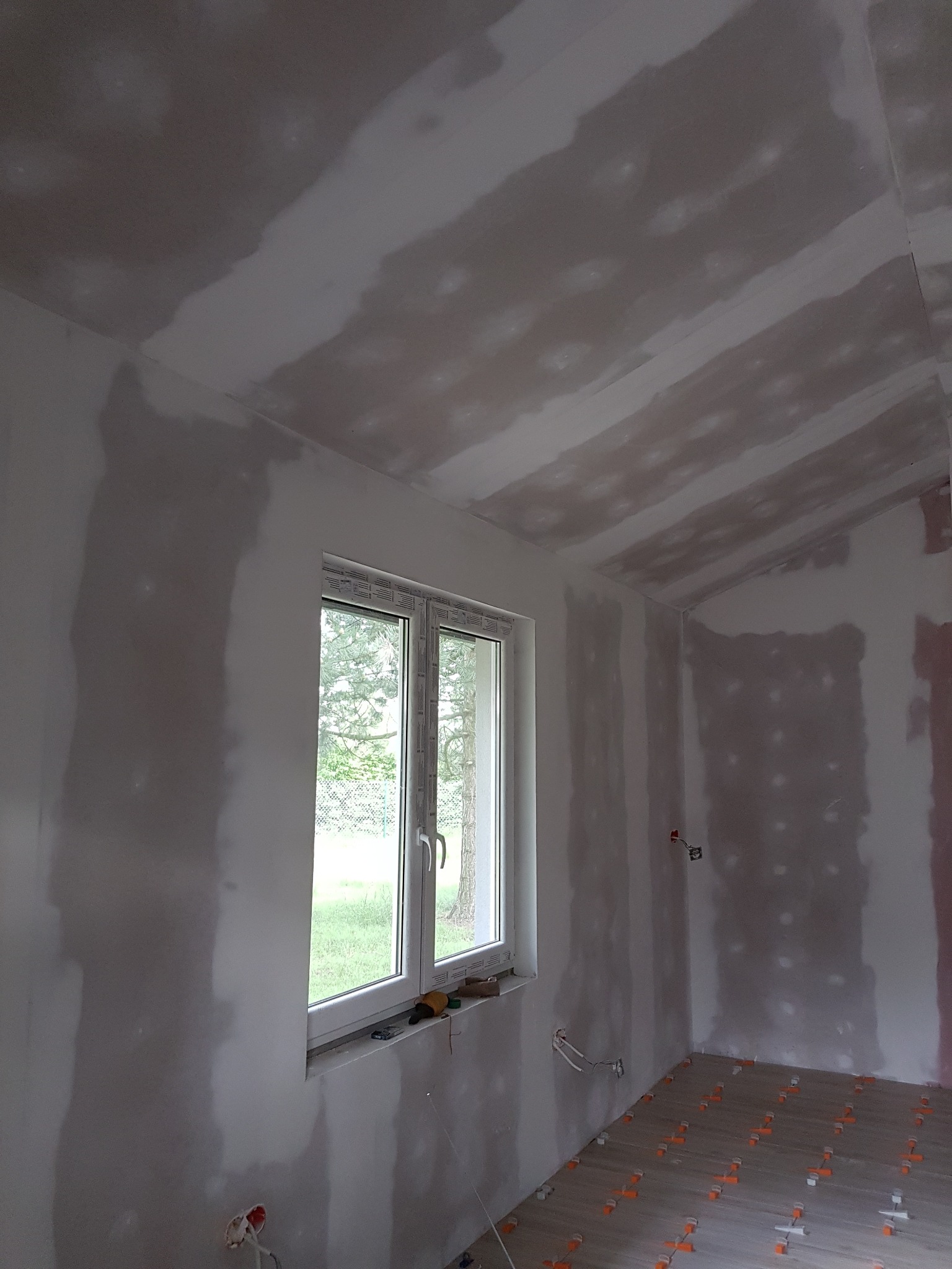 Wnętrze remontowanego pokoju z oknem: ściany i sufit pokryte warstwą gładzi szpachlowej, częściowo wyschnięte, na podłodze płytki ceramiczne z elementami poziomującymi.