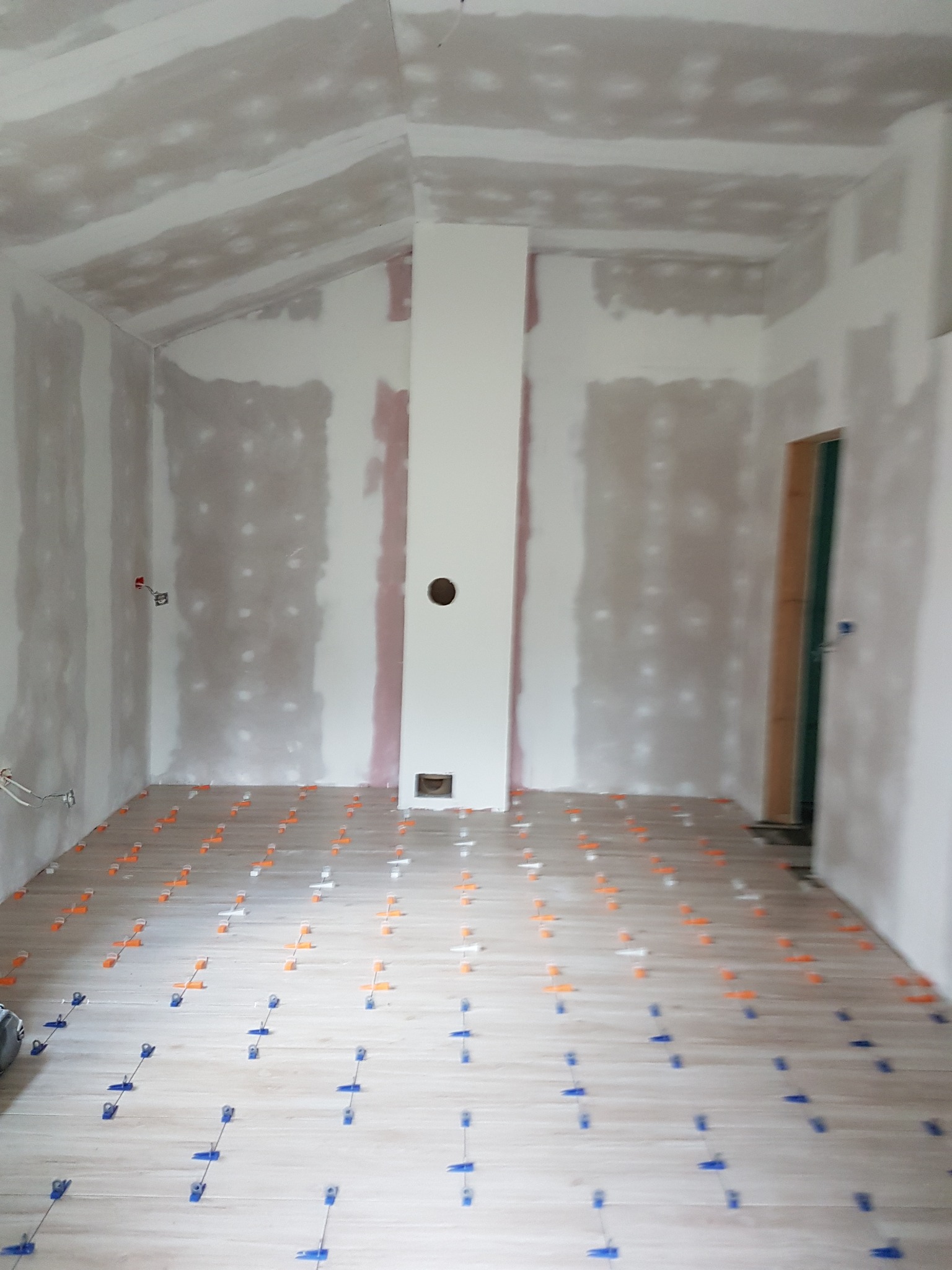 Wnętrze w trakcie prac wykończeniowych: położona jasna podłoga z systemem poziomowania płytek, ściany przygotowane pod gładzie, widoczne punkty elektryczne.