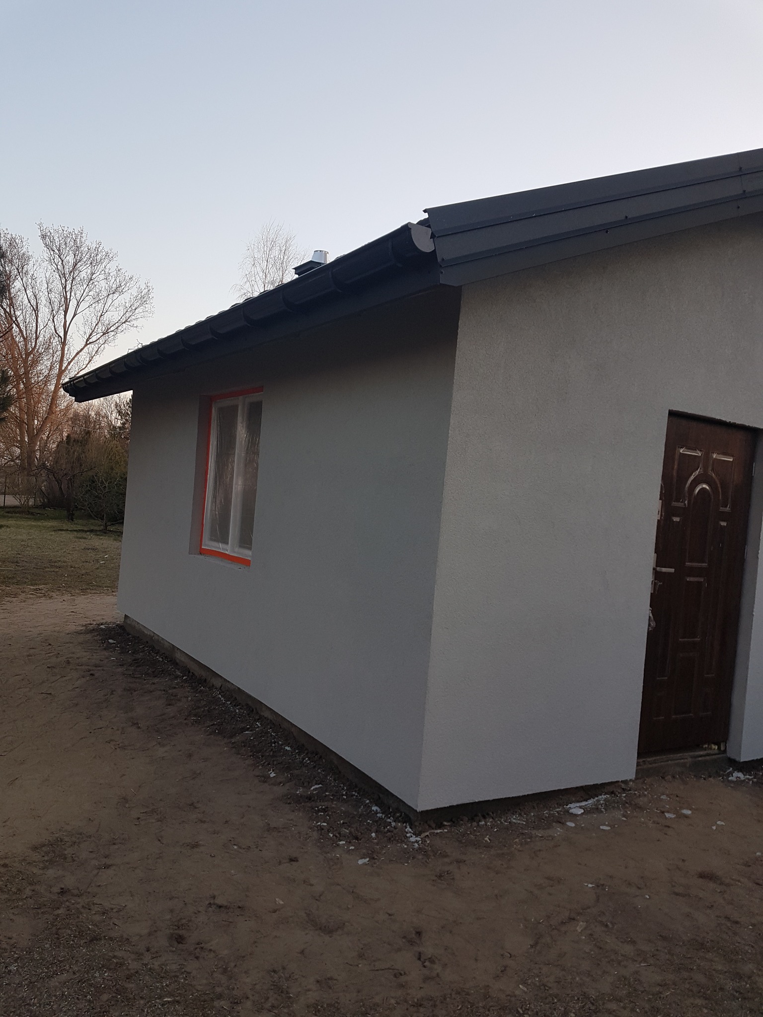 Świeżo otynkowany, szary budynek gospodarczy z brązowymi drzwiami i oknem zabezpieczonym folią malarską, widoczny wczesnym rankiem.