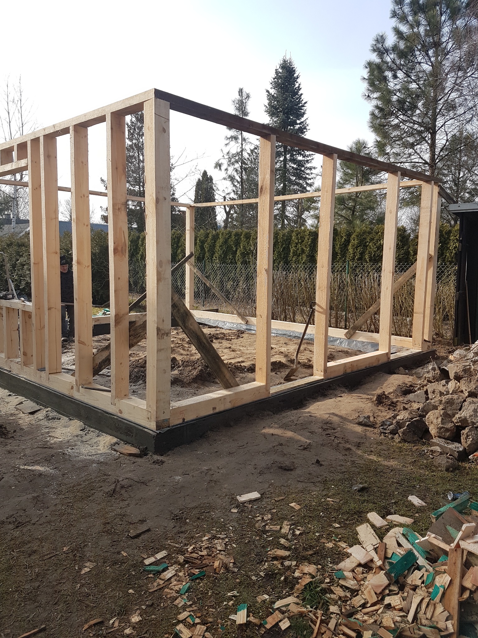 Konstrukcja szkieletowa budynku w trakcie budowy, widoczne drewniane słupy i belki, fundament z izolacją, teren budowy z piaskiem i resztkami drewna