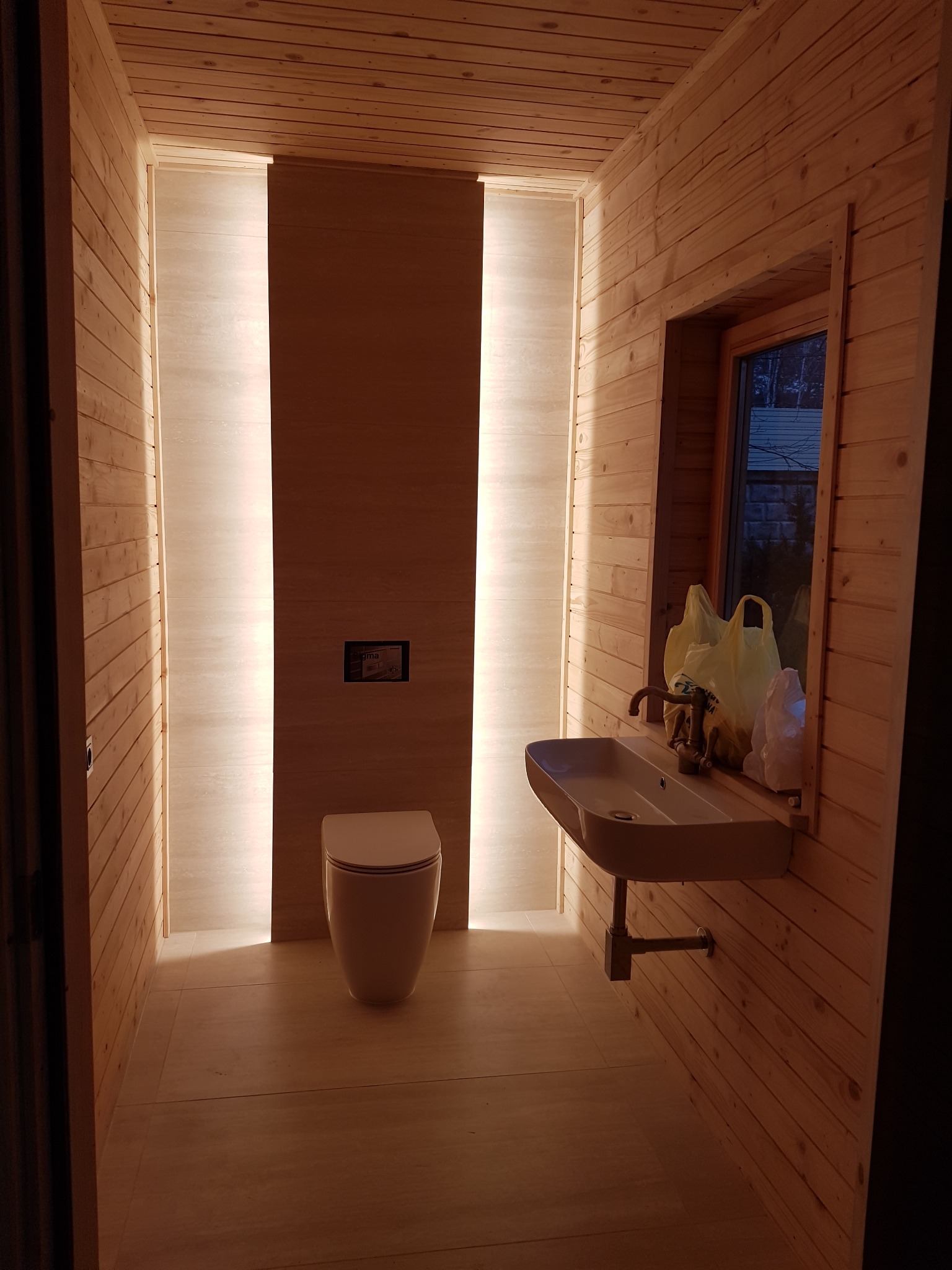 Drewniana łazienka z podświetlanymi wnękami, wiszącą toaletą i umywalką na tle okna z widokiem na zewnątrz.