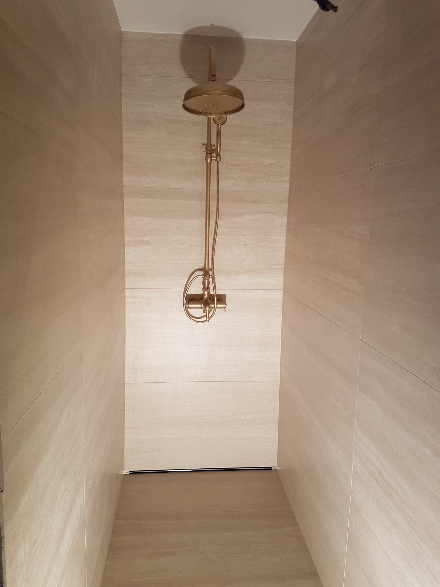 Kabina prysznicowa wyłożona beżowymi płytkami imitującymi trawertyn, z zamontowaną deszczownicą w kolorze starego złota i minimalistycznym odpływem liniowym.