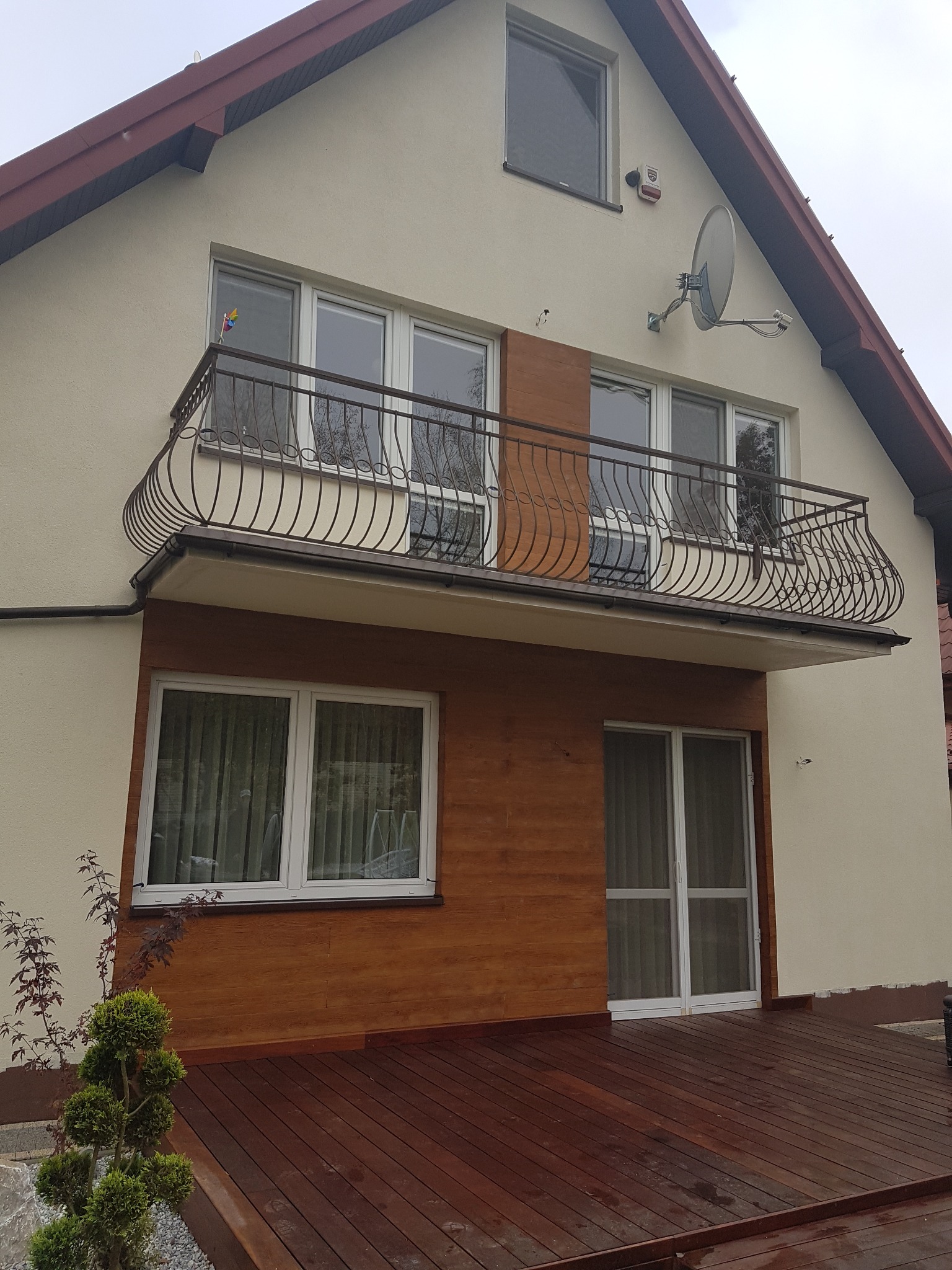 Dwukondygnacyjny dom z beżową elewacją, balkonem z ozdobną, metalową balustradą, drewnianą okładziną i tarasem z desek kompozytowych. Na ścianie widoczna antena satelitarna.