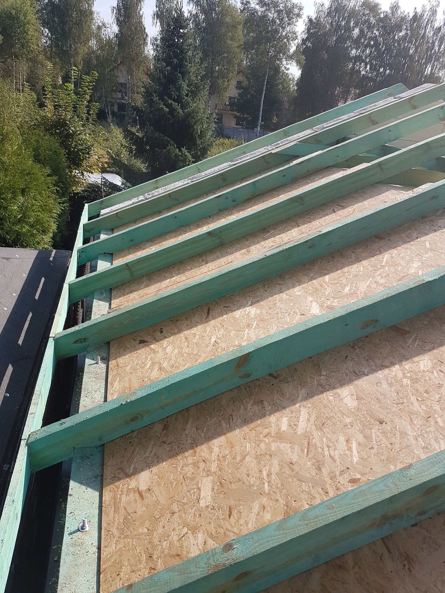 Konstrukcja dachu z belek w kolorze turkusowym i płyt OSB, widok z góry, w tle zieleń drzew.