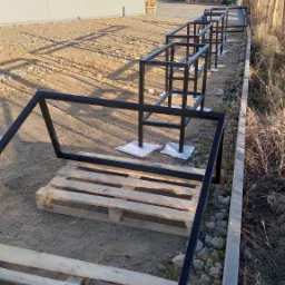 Czarne, metalowe ramy przygotowane do montażu ogrodzenia, ustawione na paletach na placu budowy, w tle budynek magazynowy i drzewa.