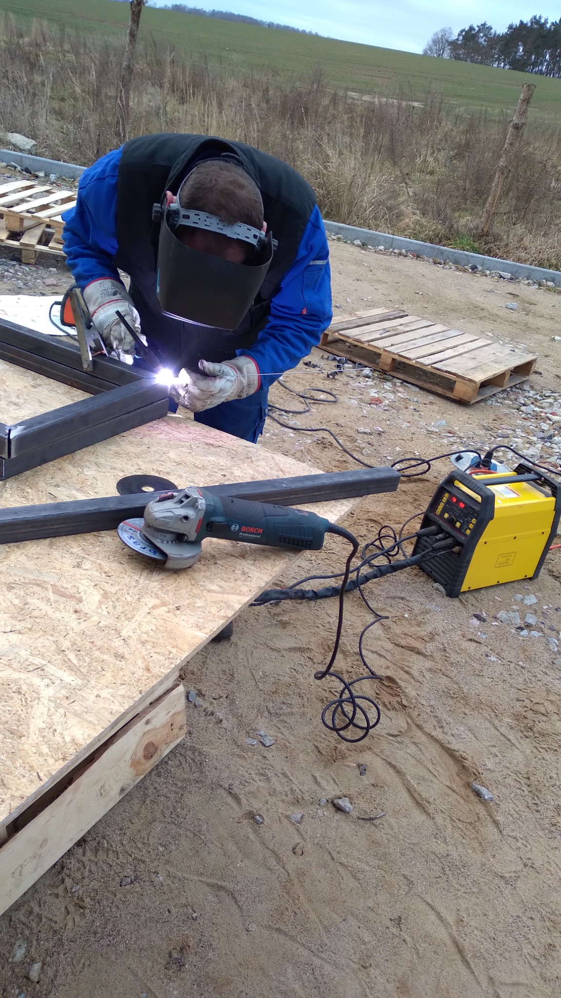 Pracownik w stroju roboczym z maską spawalniczą łączy metalowe profile spawarką na zewnątrz, obok leży szlifierka kątowa, widoczne iskry i żółta spawarka.