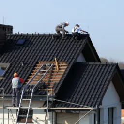 Ekipa dekarzy na dachu z czarnej dachówki, jeden pracownik na rusztowaniu przy drabinie. Część dachu bez pokrycia, widoczne łaty dachowe.