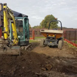 Żółto-zielona mini koparka i wywrotka na placu budowy, z wykopaną ziemią i gruzem, w tle dom jednorodzinny i ogrodzenie.