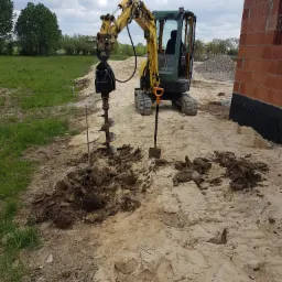 Żółto-zielona mini koparka z wiertnicą hydrauliczną wierci otwór w piaszczystej ziemi obok fundamentów budynku z czerwonej cegły, widoczna łopata i pryzma żwiru w tle.