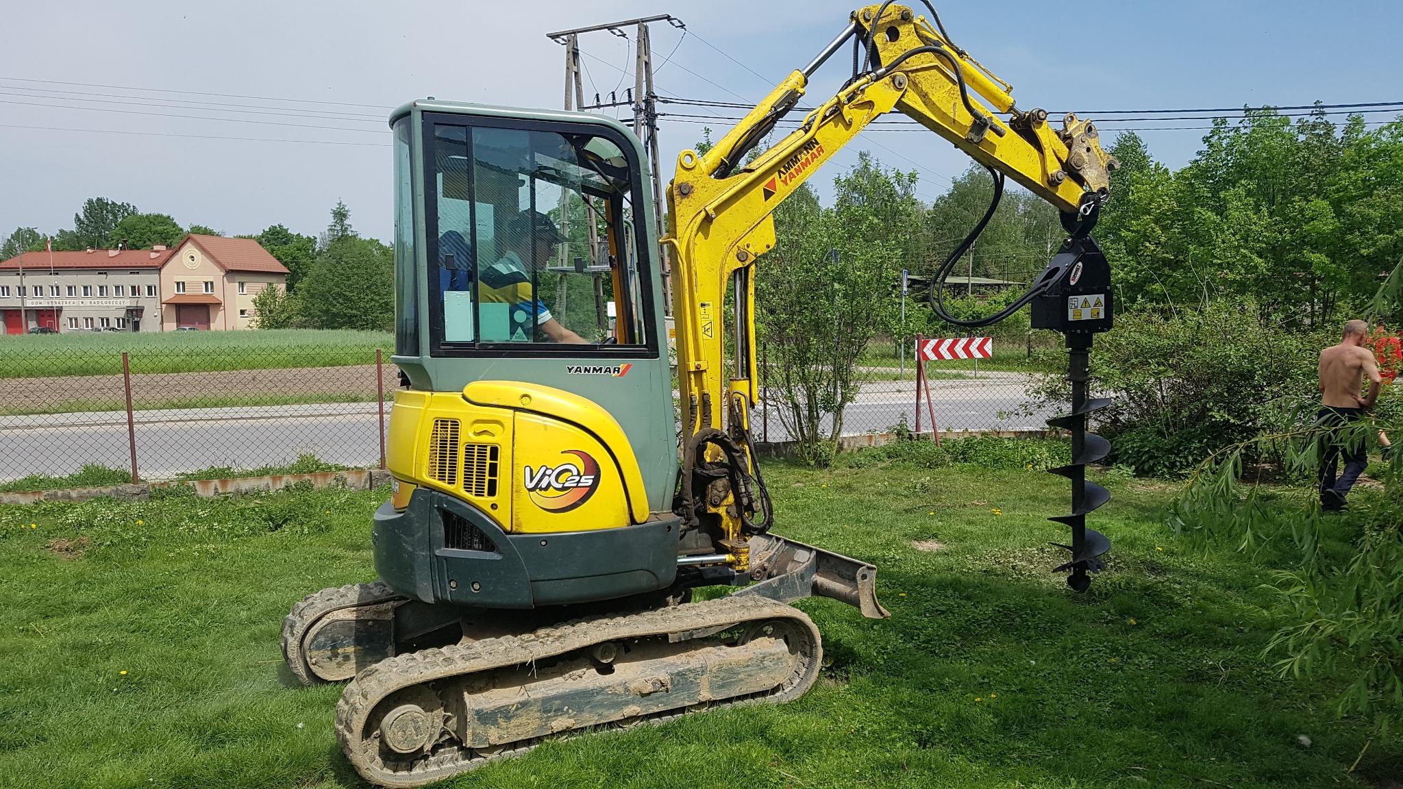 Żółto-zielona mini koparka Yanmar z wiertnicą ślimakową na trawiastym terenie, widoczny operator w kabinie, w tle budynek straży pożarnej i drzewa.