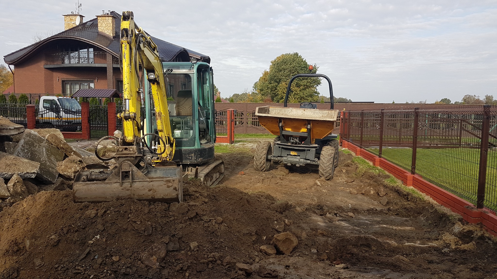 Żółto-zielona mini koparka i wywrotka na placu budowy, z wykopaną ziemią i gruzem, w tle dom jednorodzinny i ogrodzenie.