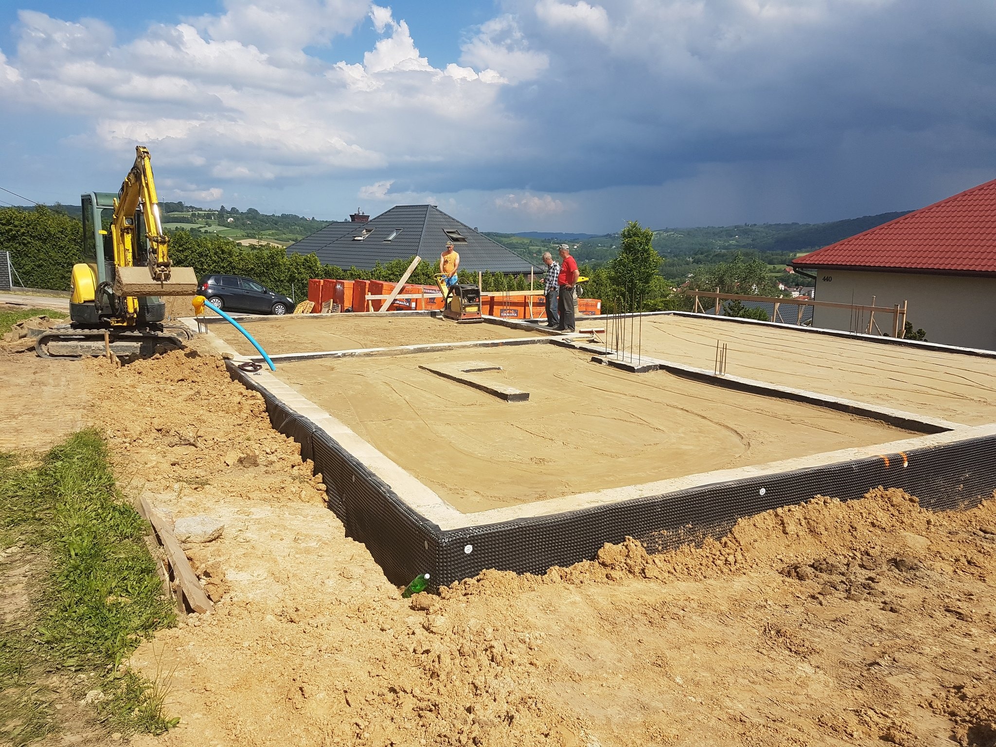 Przygotowany wykop pod fundamenty domu jednorodzinnego z izolacją, widoczny żółty mini-koparka i pracownicy, w tle krajobraz podmiejski z burzowymi chmurami.