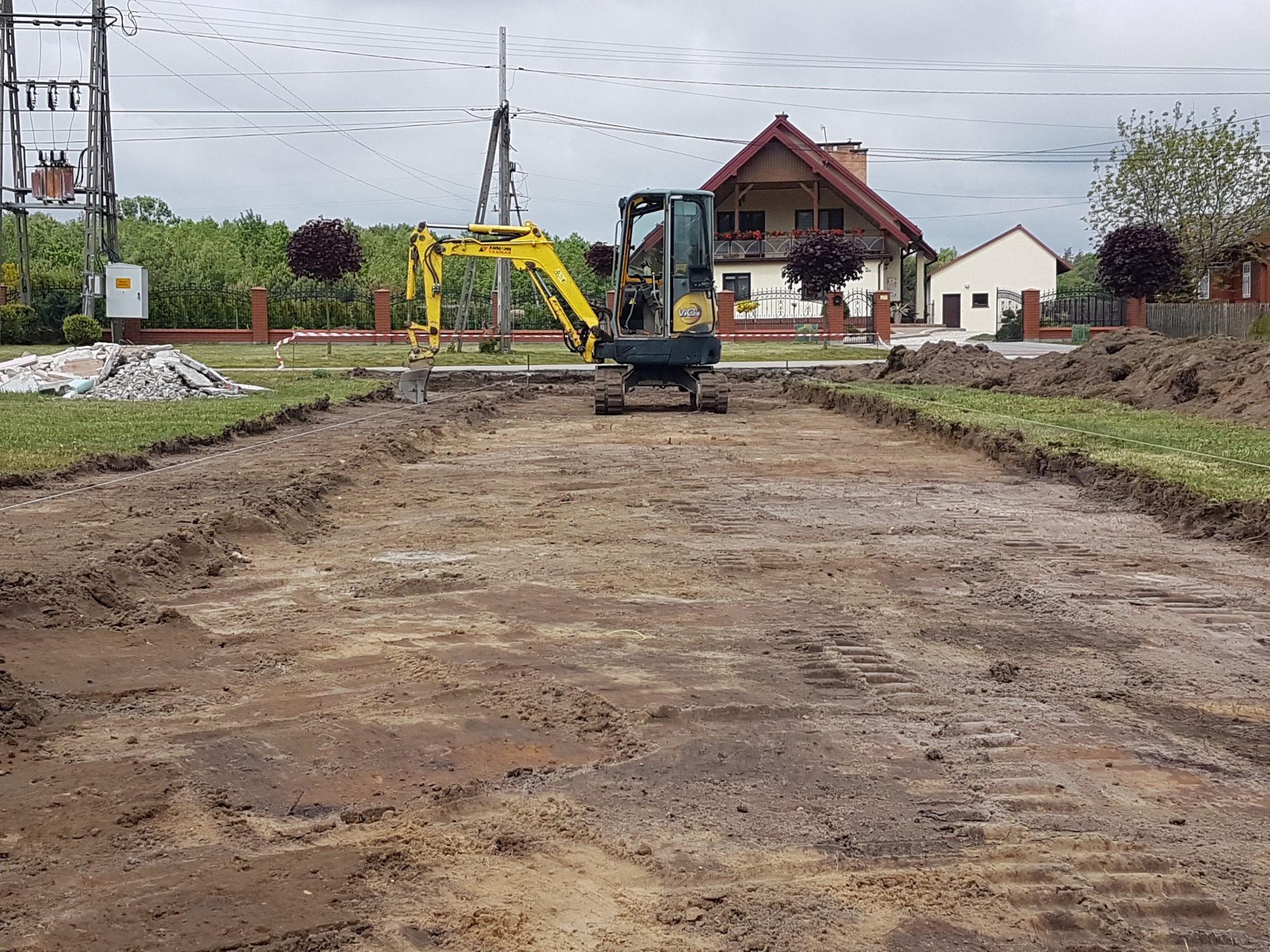 Żółta minikoparka na placu budowy, przygotowany teren pod drogę lub fundamenty, widoczne ślady gąsienic i wykopane rowy, w tle domy jednorodzinne i słup wysokiego napięcia.