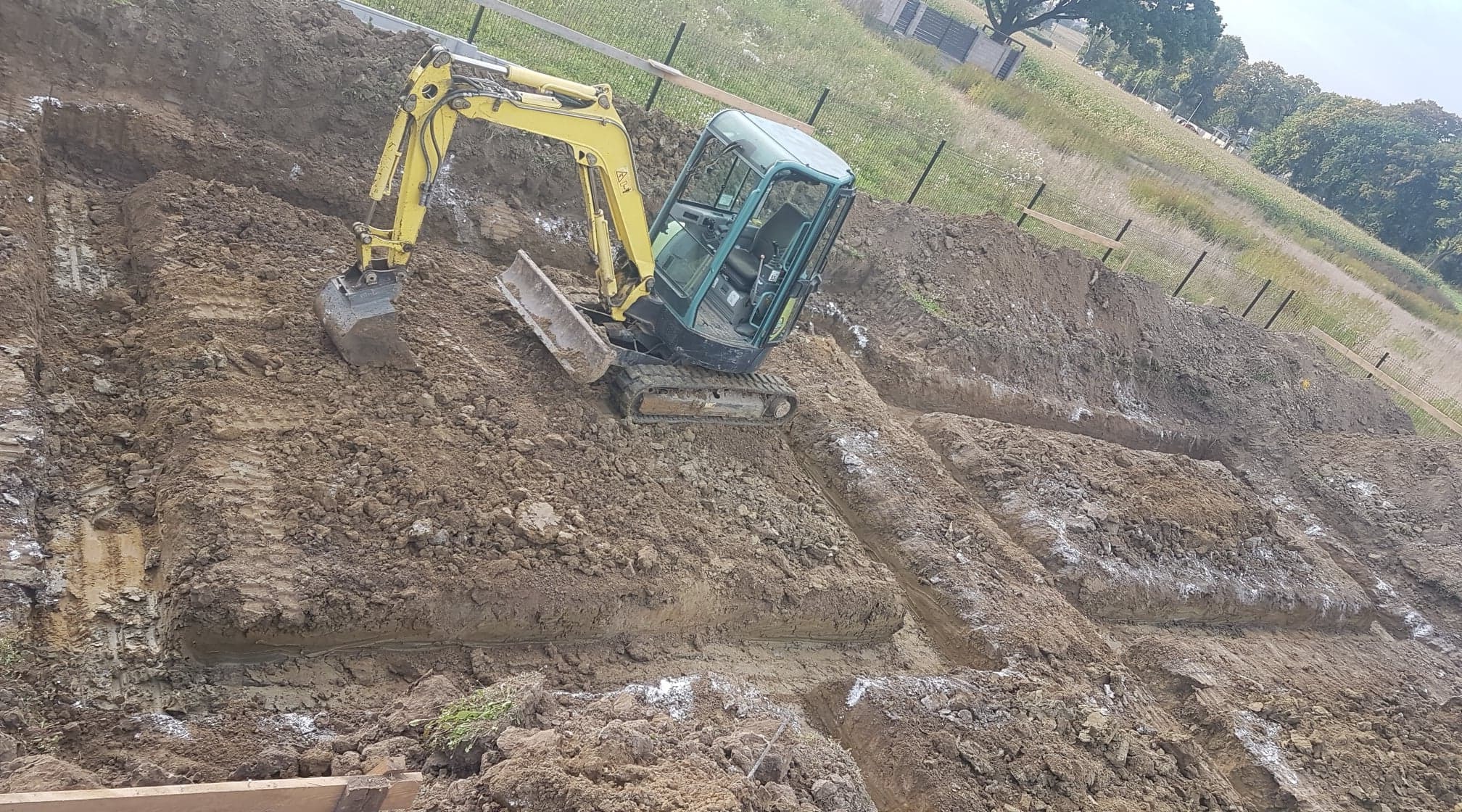Żółto-turkusowa mini koparka na gąsienicach, pracująca na zboczu, w tle zielona trawa i drzewa, widoczne wykopane rowy z białymi śladami po wapnie.