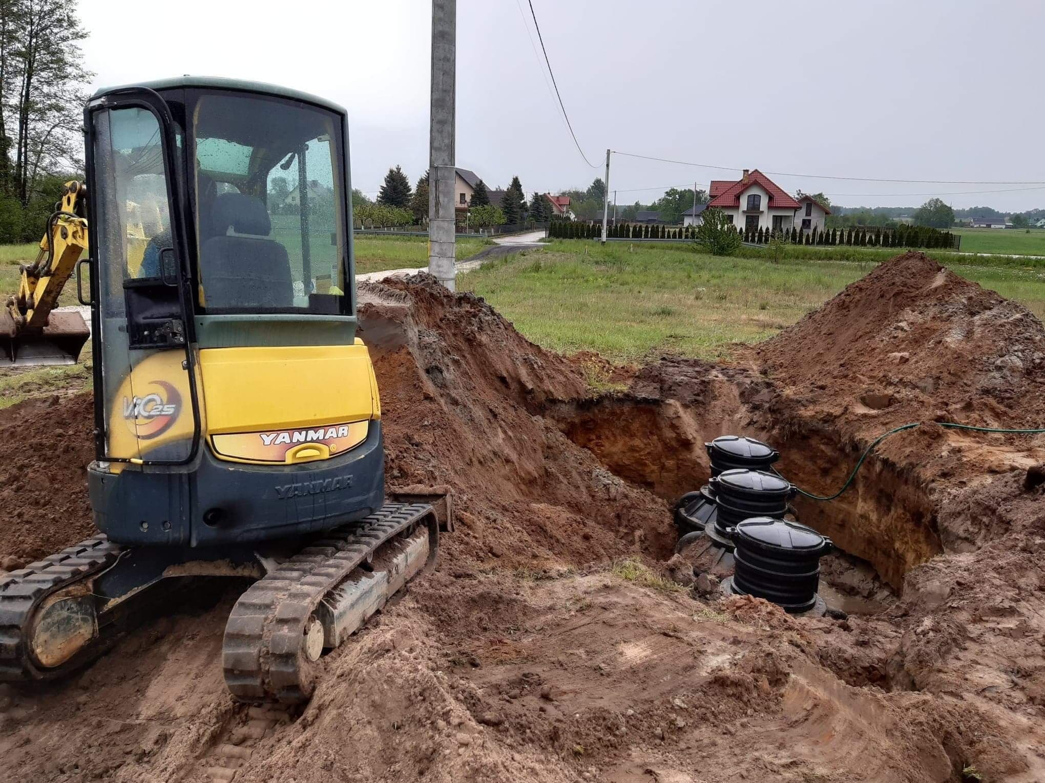Żółta mini koparka Yanmar obok wykopu z trzema czarnymi zbiornikami, widoczne świeże ślady pracy w ziemi, w tle domy i słup energetyczny.