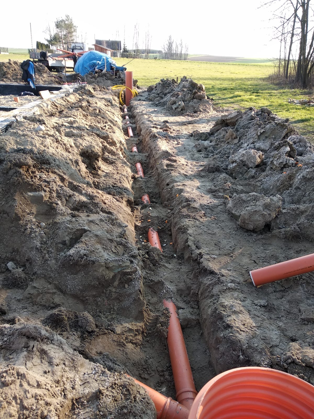 Wykopany rów z ułożonymi pomarańczowymi rurami drenażowymi, widoczne ślady opon, w tle zielone pole i budynki gospodarcze.