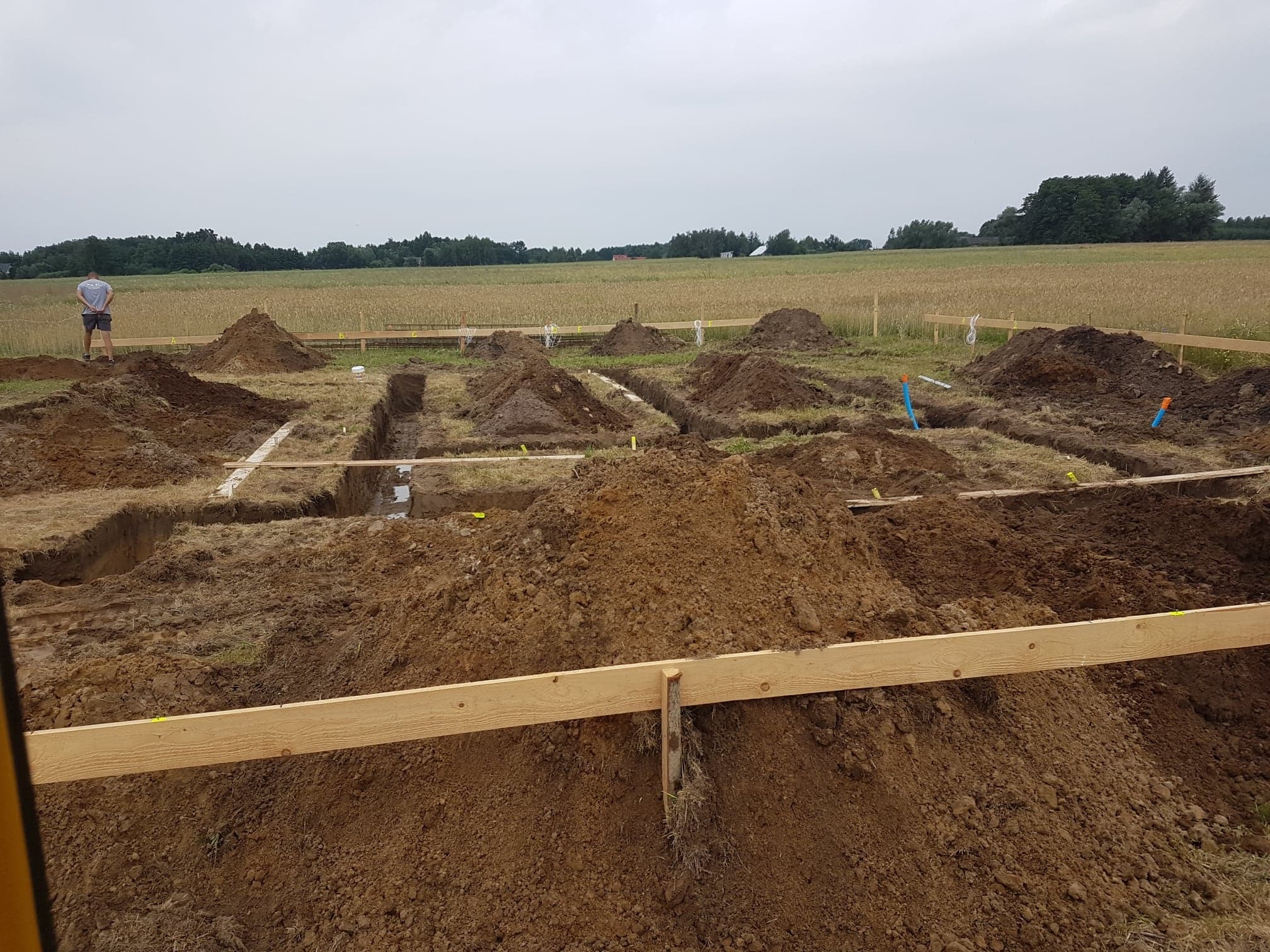 Przygotowanie terenu pod budowę w Tarnowie: wykopane rowy pod fundamenty z drewnianym szalunkiem, widoczny mężczyzna w tle i pole uprawne.