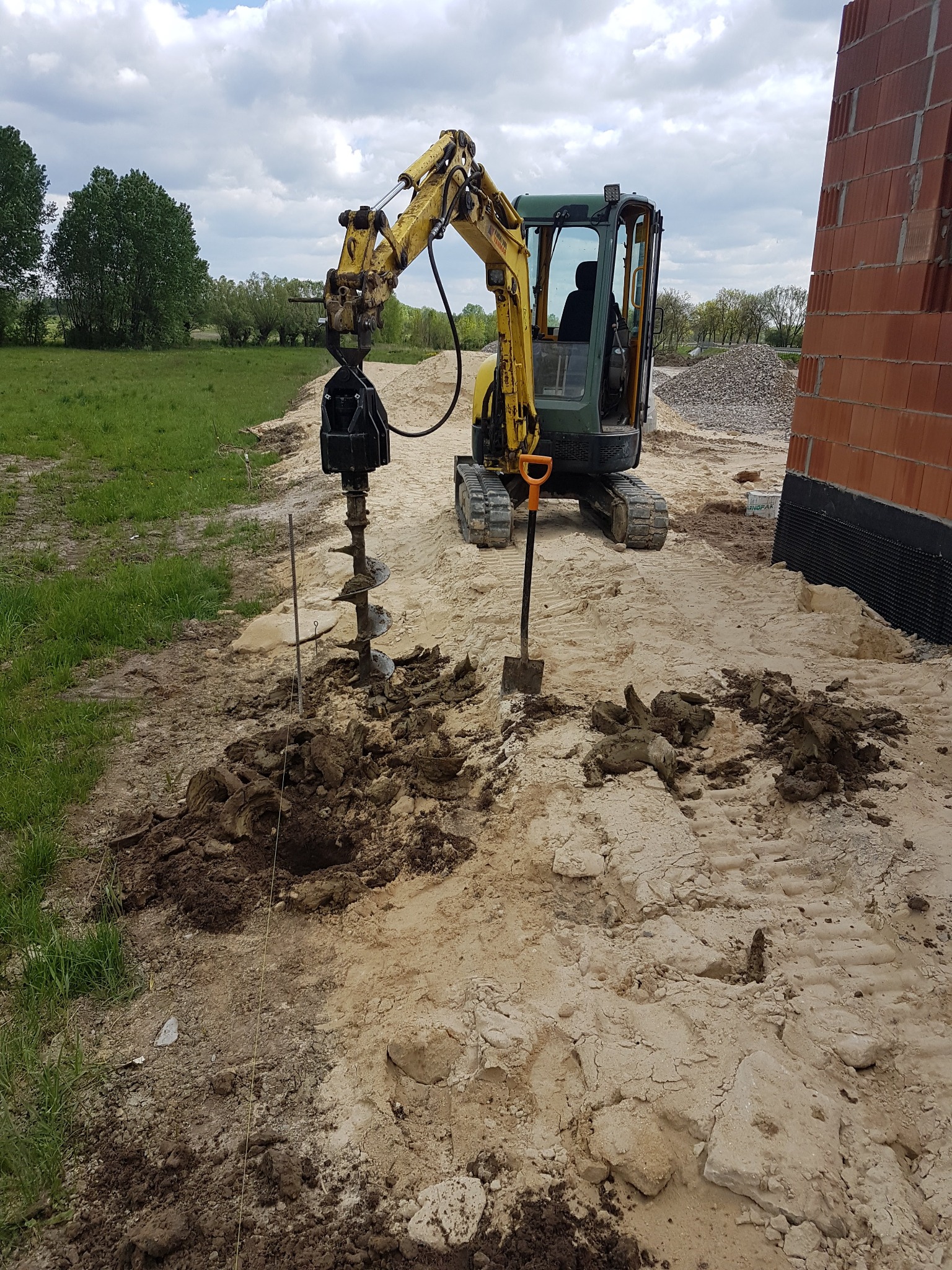 Żółto-zielona mini koparka z wiertnicą hydrauliczną wierci otwór w piaszczystej ziemi obok fundamentów budynku z czerwonej cegły, widoczna łopata i pryzma żwiru w tle.