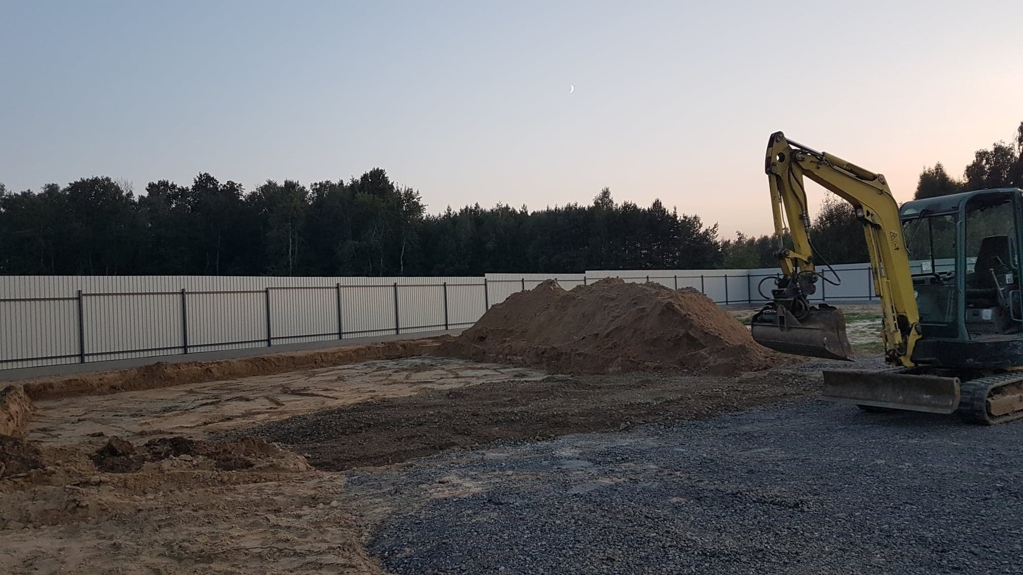 Żółta mini koparka na placu budowy, świeżo wykopane fundamenty, sterta ziemi i metalowe ogrodzenie w tle, widoczne wczesnym rankiem przy księżycu.