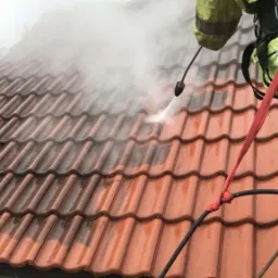 Pracownik w kombinezonie i uprzęży, z bliska myje dachówkę ciśnieniową myjką, widoczna różnica w czystości pomiędzy umytą a brudną częścią dachu.