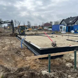 Przygotowany szalunek z izolacją pionową fundamentów pod budowę domu jednorodzinnego, zagęszczarka wibracyjna na piasku, w tle koparki Volvo i kontenery budowlane.