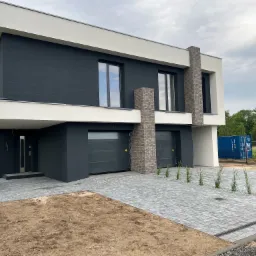 Nowoczesny dom murowany z ciemnoszarą elewacją i białymi elementami, kostka brukowa na podjeździe, widoczny kontener budowlany w tle.