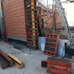 Wznoszenie ściany z czerwonej cegły ceramicznej, wzmocnionej zbrojeniem i szalunkiem, widoczne elementy konstrukcyjne i drabina na placu budowy.