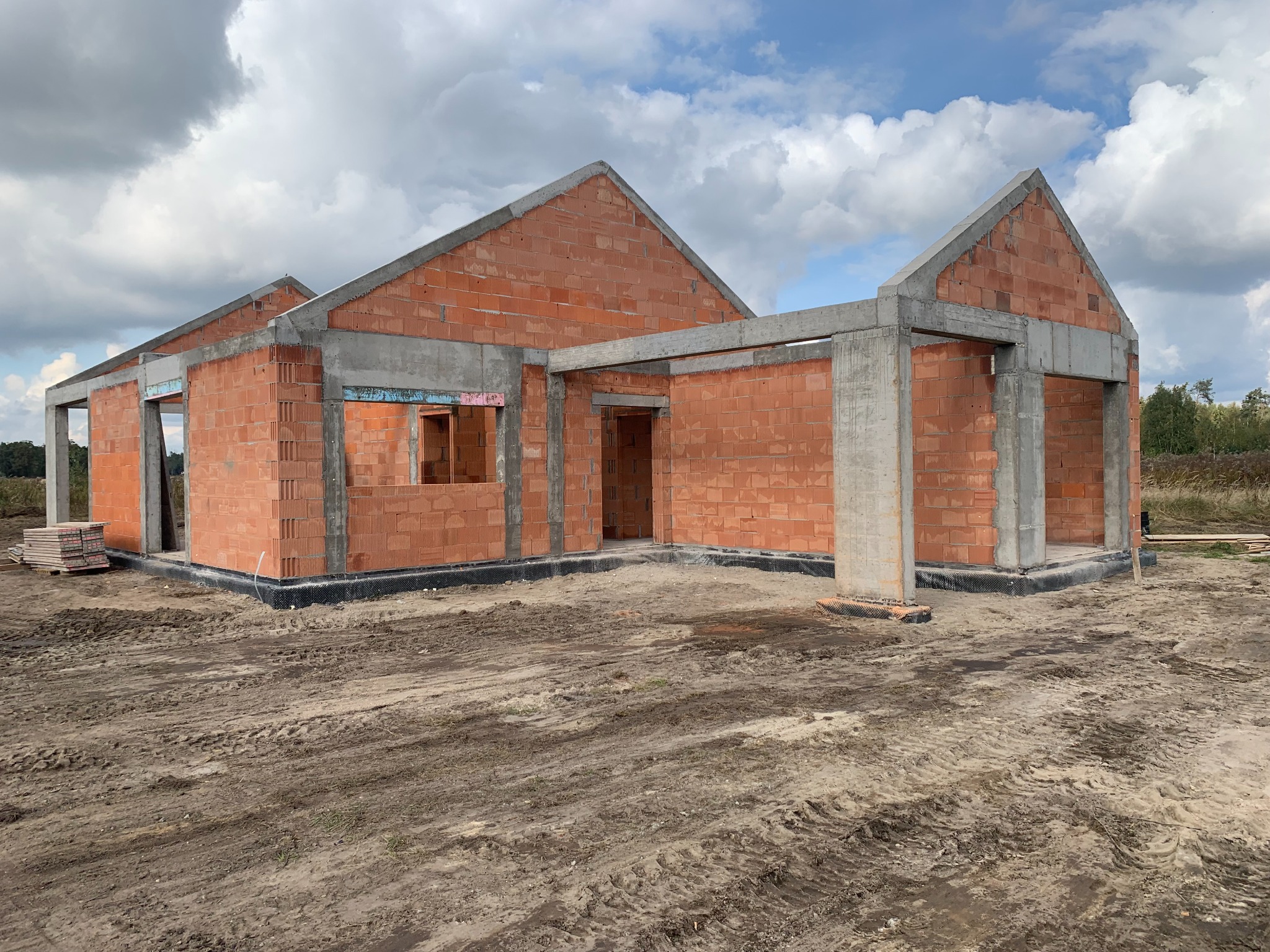 Niedokończony budynek z czerwonej cegły z betonowymi elementami konstrukcyjnymi pod niebem z chmurami, widoczne ślady opon na ziemi.