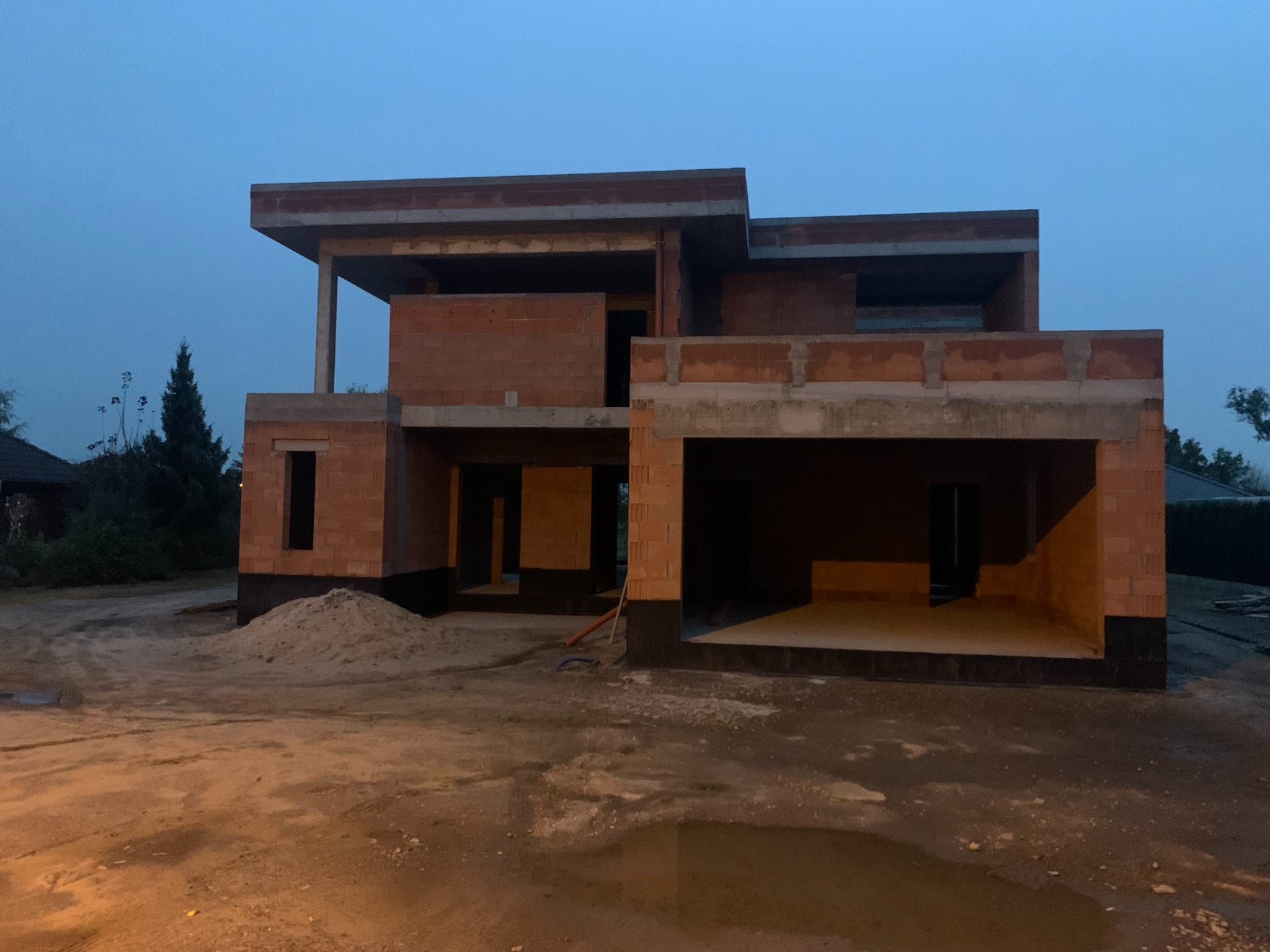 Surowy stan dwukondygnacyjnego domu z pustaków ceramicznych, z widoczną konstrukcją żelbetową, bez okien i drzwi, na tle pochmurnego nieba o zmierzchu.